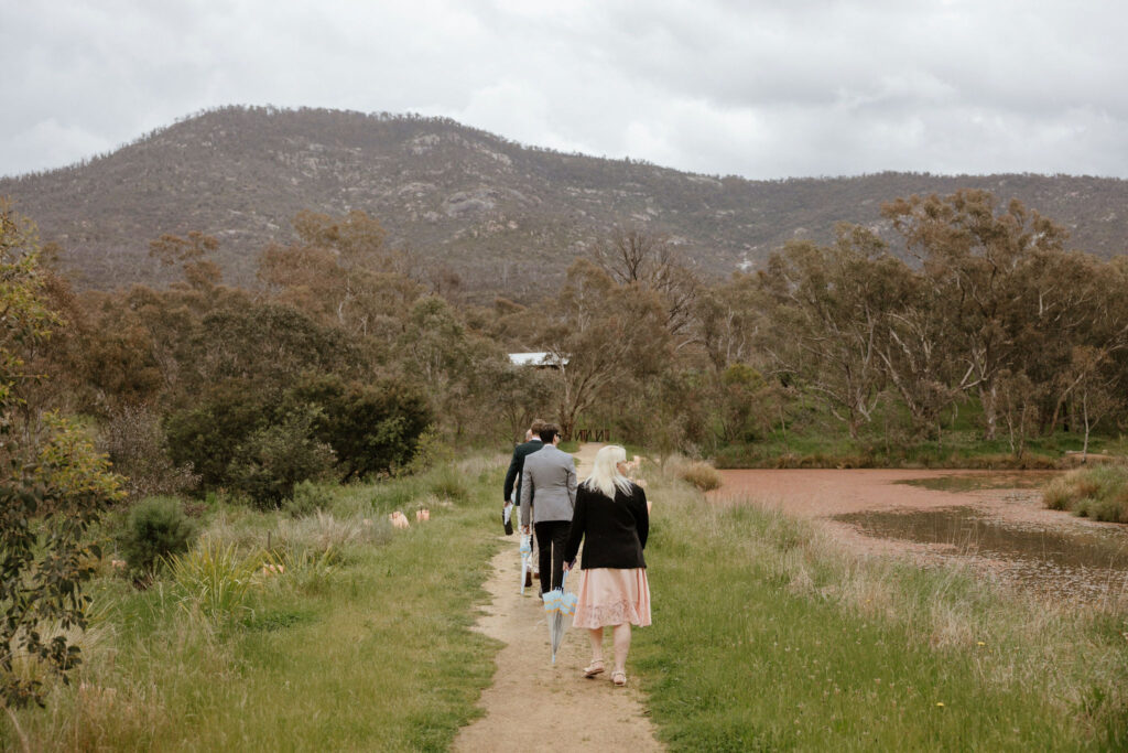 A small group of family and best friends walk under umbrellas along a dirty path in a national forest to find an elopement ceremony spot. Mt Tennent is in the distance.