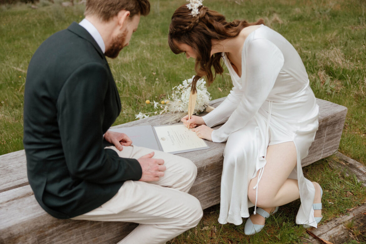 A bride and groom sit on a log to sign their wedding certificate