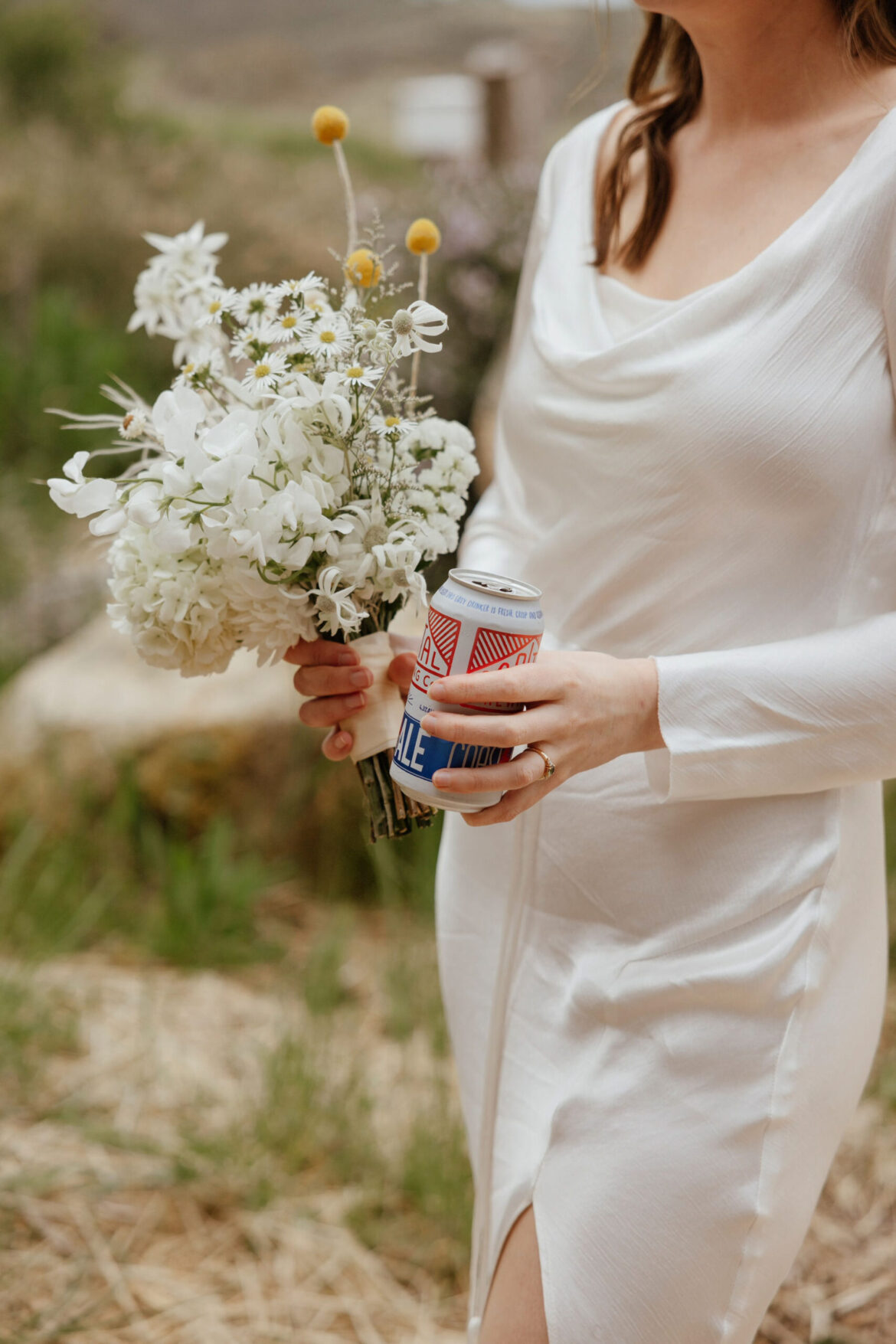 A bride holds both a beer and some wedding flowers on her wedding day