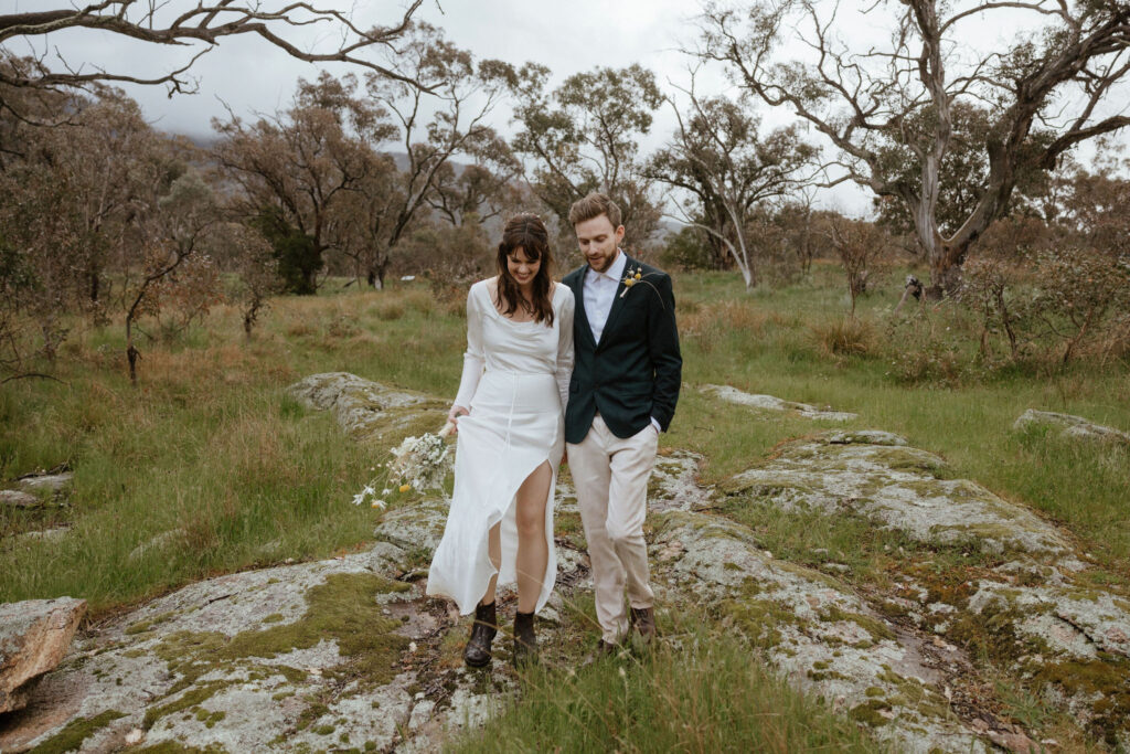 A groom wearing a suit walks with his bride wearing a long silky dress and blundstone boots, through a rocky outcrop in the bush