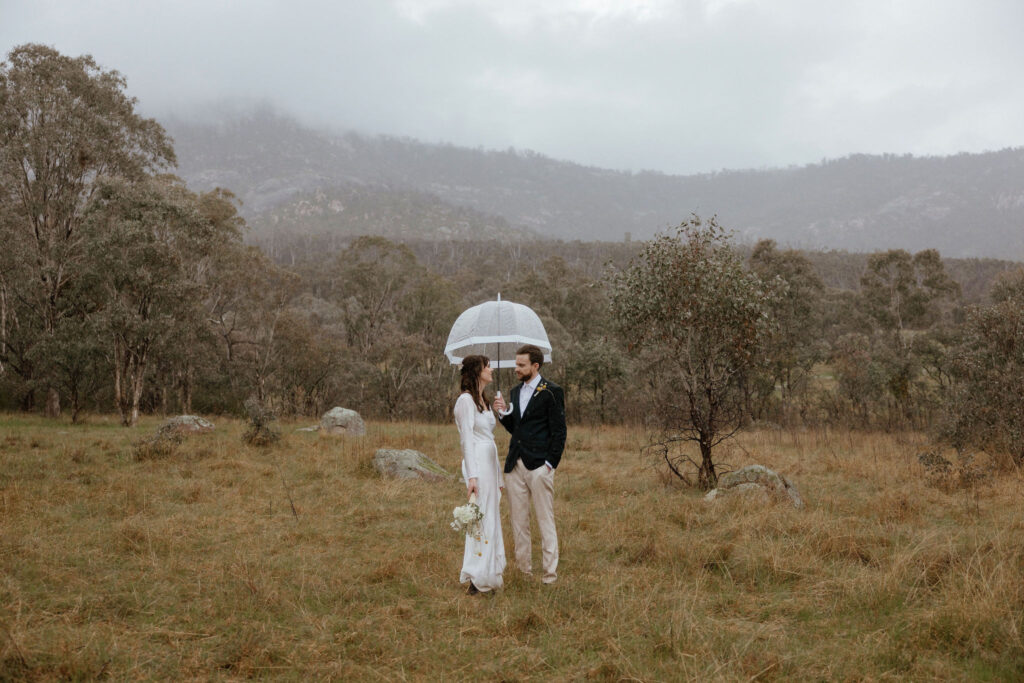 A bride and groom cuddle under an umbrella while it rains, with Mt Tennent in the background