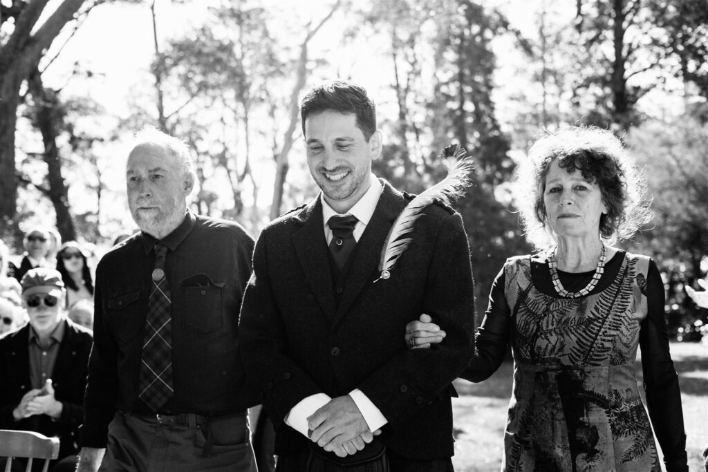 A groom wearing a kilt walks down the aisle in the arms of both his parents