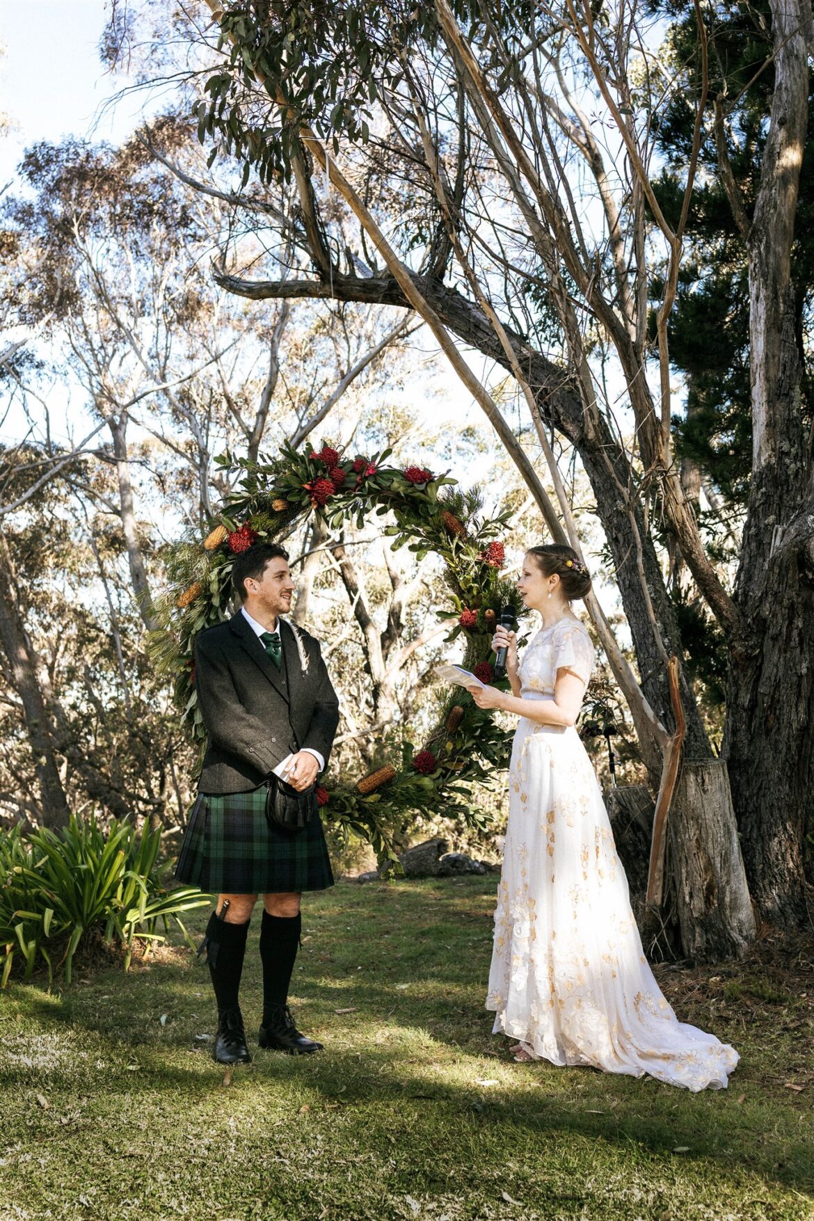 A groom wearing a kilt and a bride wearing a vintage wedding dress both read their vows to each other in their wedding ceremony in the Blue Mountains