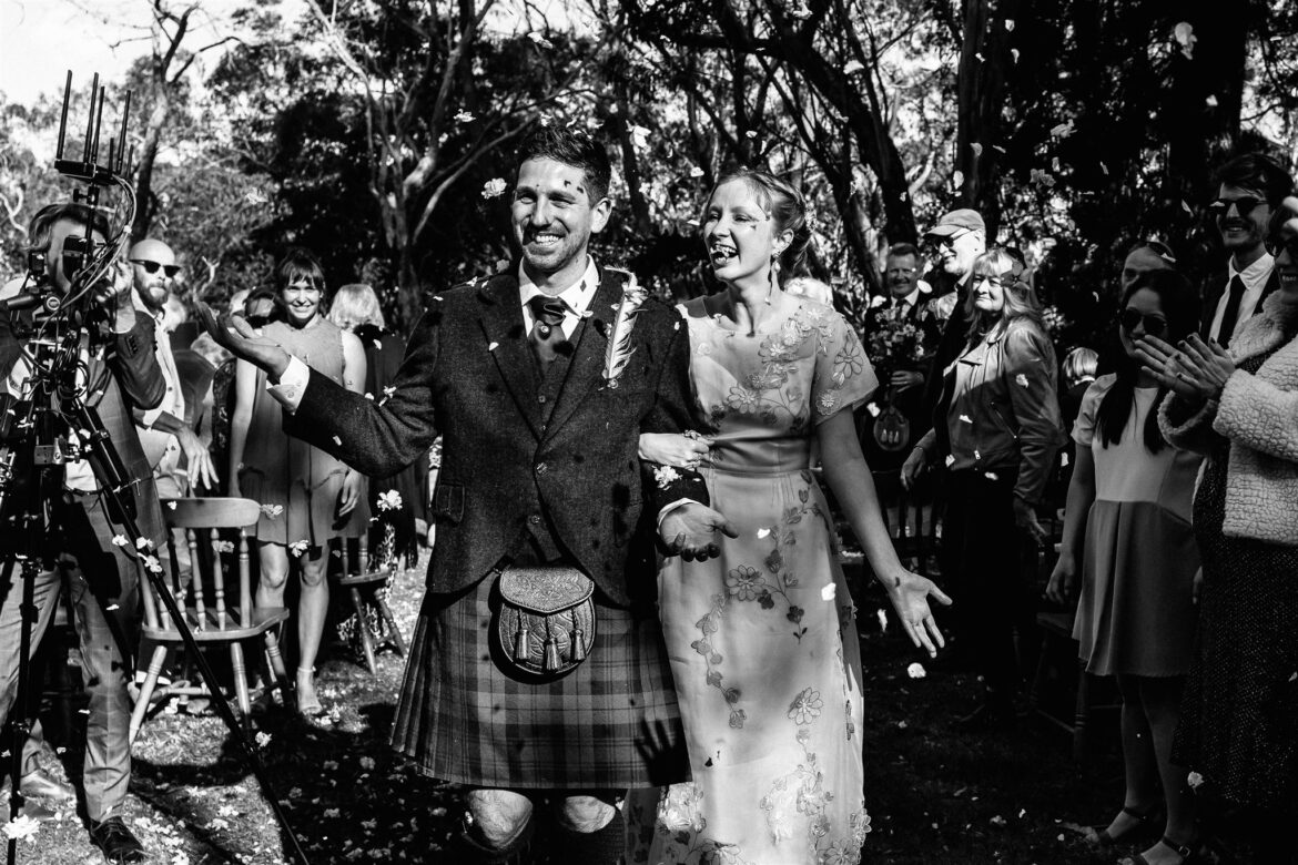 A groom wearing a kilt and a bride wearing a vintage wedding dress both exit their wedding ceremony with leaf confetti being thrown on them in celebration