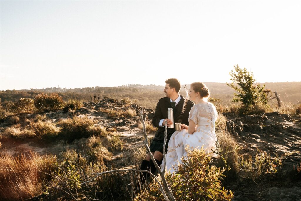 A bride wearing a vintage wedding dress and a groom wearing a kilt, both share a cup of tea sitting on rocks at the Blue Mountains, with personalised mugs