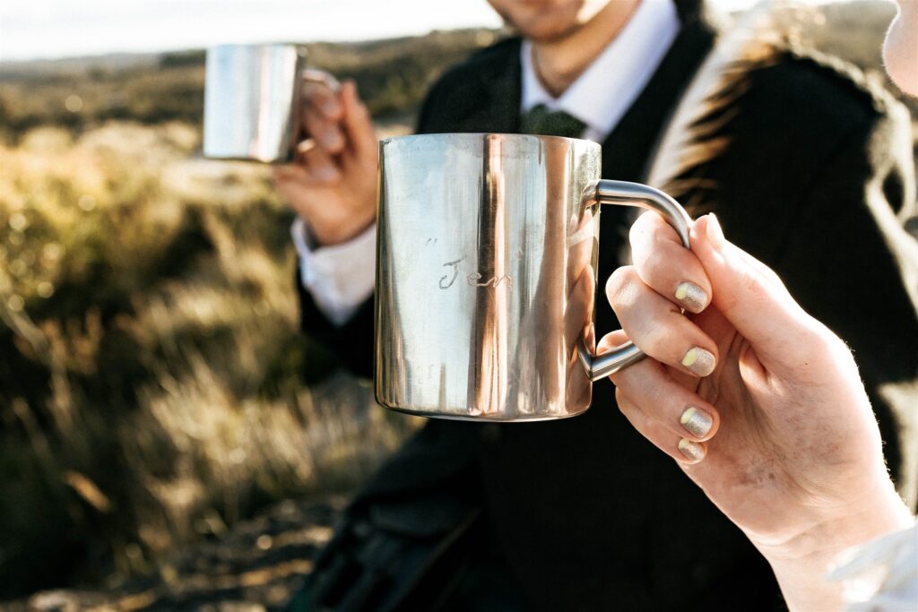 A bride wearing a vintage wedding dress and a groom wearing a kilt, both share a cup of tea sitting on the mountain, with personalised mugs