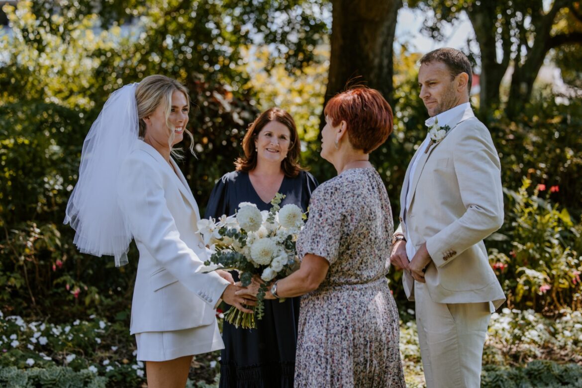 A bride wearing a white suit and skirt with a veil and groom wearing an offwhite suit, stand in a garden at a ceremony with a celebrant and a parent saying kind words to the bride.