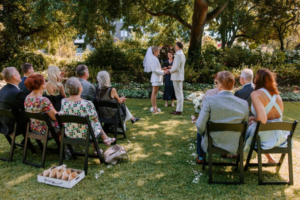 A wedding ceremony takes place in the gardens of Highwic in Newmarket - Central Auckland. A bride wearing a suit jacket and white skirt and veil holds the hands of the groom wearing an offwhite suit. The celebrant and guests watch.