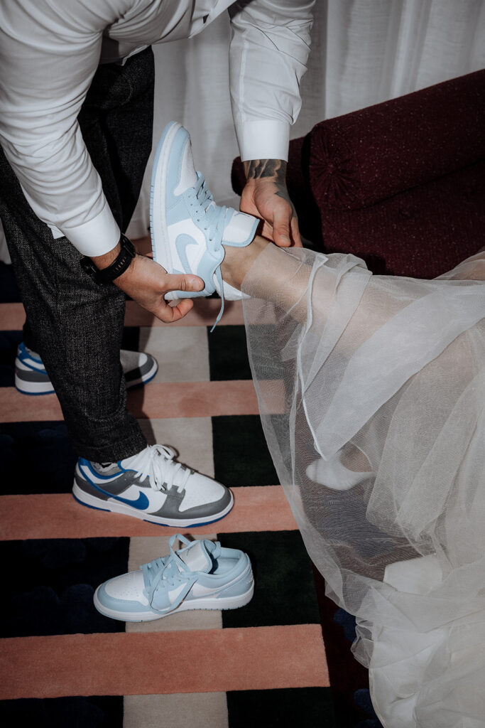 A groom helps put matching Nike sneakers on his bride. She is wearing a big white dress. 