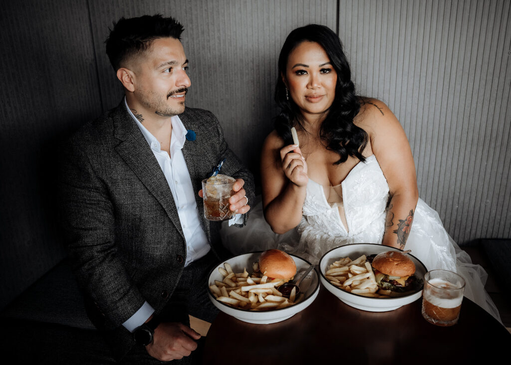 A bride and groom are dressed glamorously in wedding clothes but sit in a bar eating burgers and fries