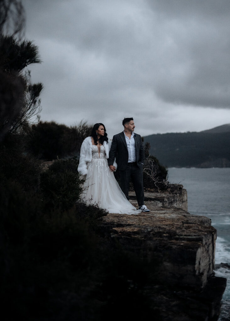 A bride and groom hold hands on a stormy coastline of Hobart, Tasmania
