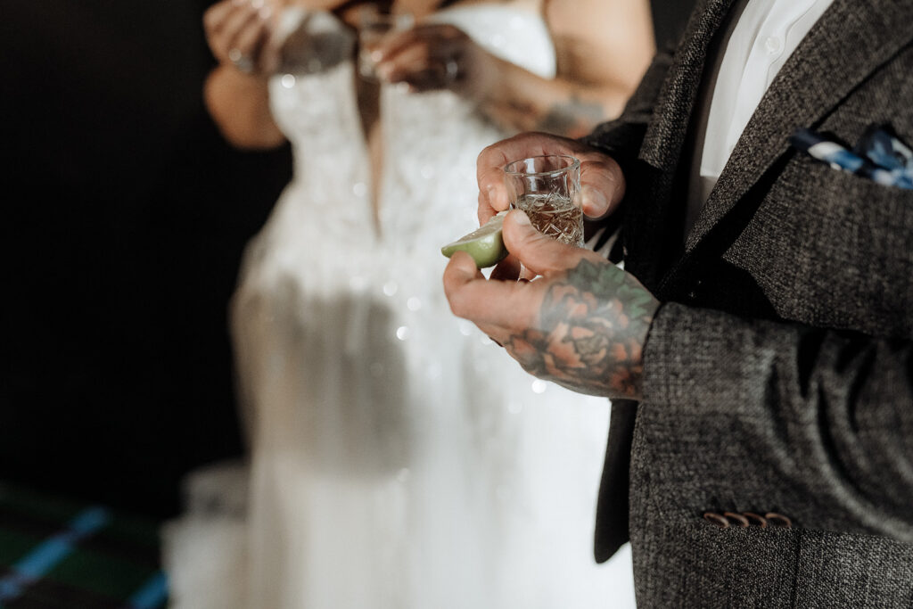 A bride and groom wearing a white wedding dress and veil and a grey suit, both drink tequila shots at their wedding ceremony