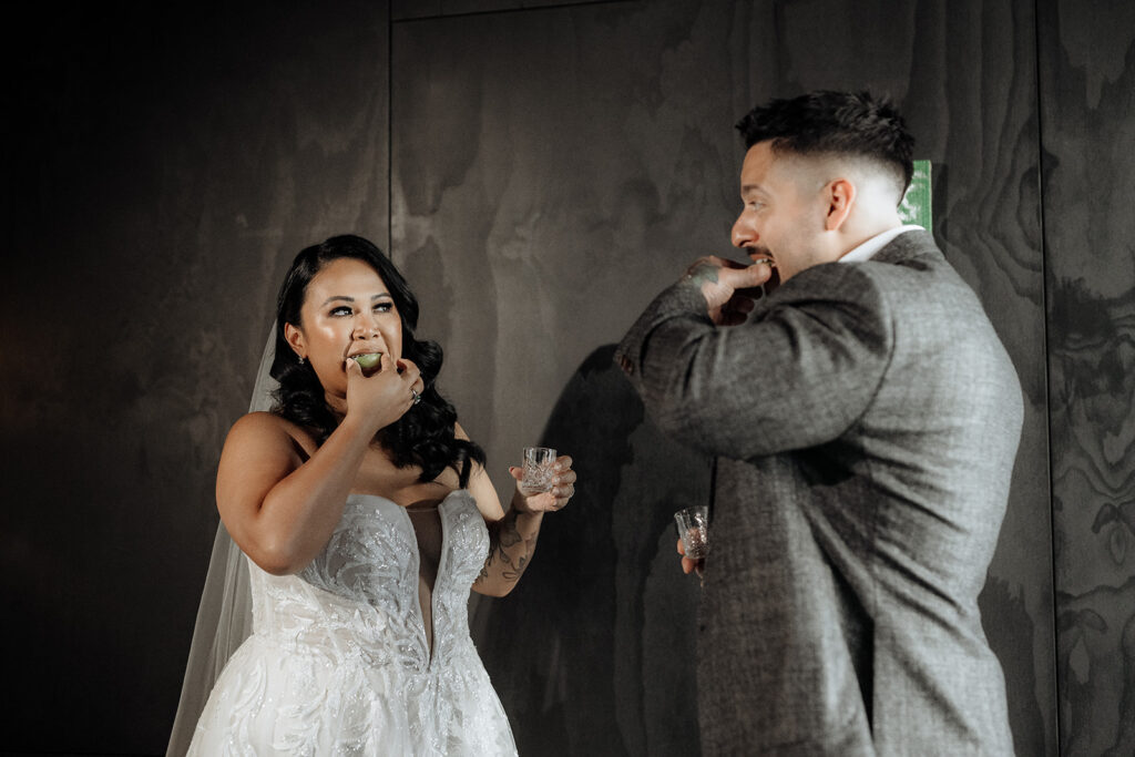 A bride and groom wearing a white wedding dress and veil and a grey suit, both drink tequila shots at their wedding ceremony